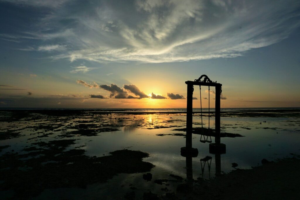 Silhueta de uma pessoa em pé sobre uma formação rochosa perto de um corpo d'água durante o pôr do sol