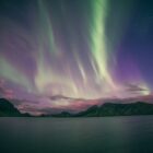 Green and purple aurora borealis above a dark lake and distant mountain range