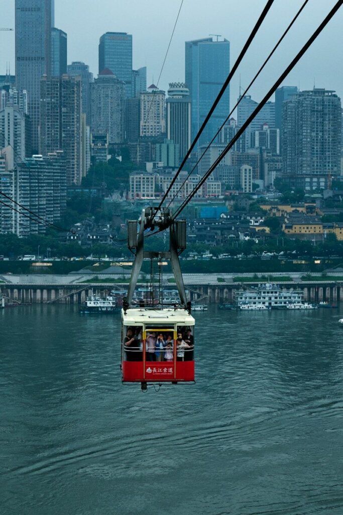 Red cable car crosses river, towering city skyline in background