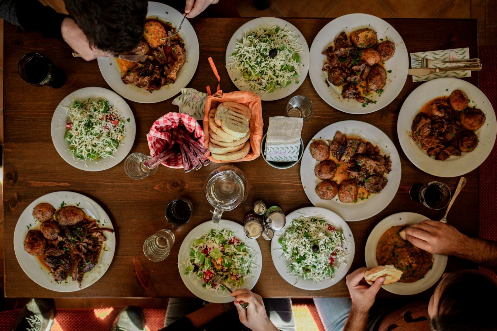 Overhead view of a wooden table covered with shared plates of food and hands reaching in
